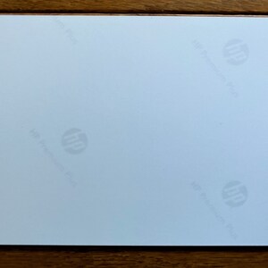 May include: A rectangular sheet of white photo paper with a smooth finish. The back side of the paper features the HP logo and the text HP Premium Plus printed in a light gray, repeating pattern across the surface of the white paper sheet.