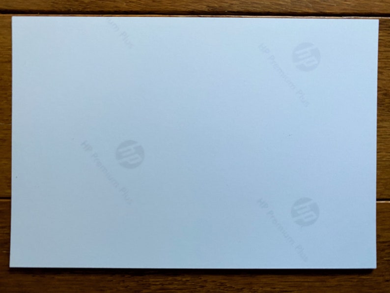May include: A rectangular sheet of white photo paper with a smooth finish. The back side of the paper features the HP logo and the text HP Premium Plus printed in a light gray, repeating pattern across the surface of the white paper sheet.