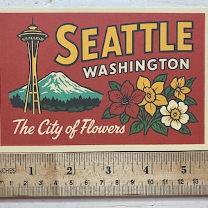May include: A rectangular postcard with a red background featuring the Space Needle, a mountain, and flowers. Text reads SEATTLE WASHINGTON and The City of Flowers. A wooden ruler shows the card is approximately 6 inches wide by 4 inches tall.