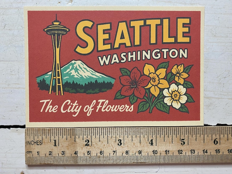 May include: A rectangular postcard with a red background featuring the Space Needle, a mountain, and flowers. Text reads SEATTLE WASHINGTON and The City of Flowers. A wooden ruler shows the card is approximately 6 inches wide by 4 inches tall.