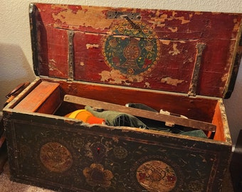 May include: An antique wooden chest with a hinged lid, painted in dark green and red with floral and vase motifs. The chest is open, revealing the interior and contents, including a small orange box and a wooden bar.