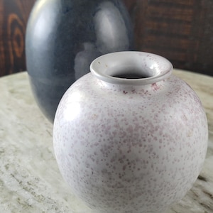 May include: A small, spherical white vase with a speckled pink pattern sits in the foreground. Behind it stands a larger, smooth, dark blue-gray oval vase. Both ceramic vessels feature a simple, rounded opening at the top for floral arrangements.