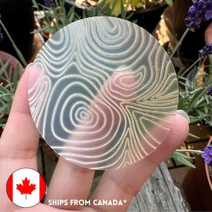 May include: A clear, circular sticker with a topographic map design in gold and white. The sticker is held in a hand, with a Canadian flag in the lower left corner. The sticker has a detailed, swirling pattern.
