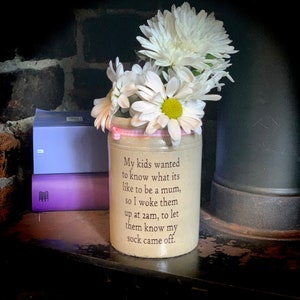 May include: A white ceramic jar with a pink ribbon around the top. The jar has a quote printed on it that reads: "My kids wanted to know what it's like to be a mum, so I woke them up at 2am, to let them know my sock came off."