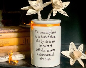 May include: A white ceramic cylindrical jar with an orange ribbon tied around the rim. The jar features black printed text that reads: You normally have to be bashed about a bit by life to see the point of daffodils, sunsets and uneventful nice days.