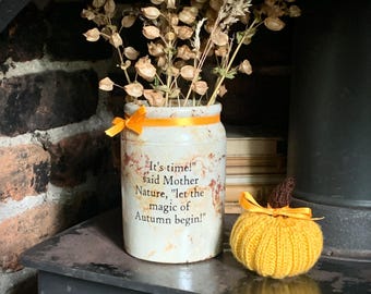 May include: A cream-colored ceramic jar with a distressed finish, tied with an orange ribbon, featuring the text "It's time!" said Mother Nature, "let the magic of Autumn begin!" and filled with dried seed pods, alongside a small, knitted yellow pumpkin.
