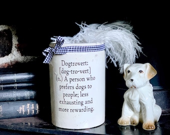 May include: A white ceramic cylindrical crock featuring a blue and white gingham ribbon tied around the top. The text reads: Dogtrovert: [dog-tro-vert] (n.) A person who prefers dogs to people; less exhausting and more rewarding. A white dog figurine sits beside it.