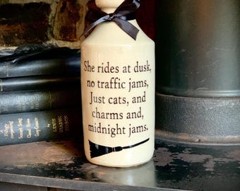 May include: A cream-colored ceramic bottle with a black ribbon tied around the neck. The bottle features black text that reads: She rides at dusk, no traffic jams, Just cats, and charms and, midnight jams. A black graphic of a broom is below the text.
