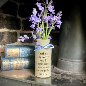 May include: A cream-coloured vase displays a bunch of purple bluebell flowers. The vase is adorned with a blue and white ribbon and the quote "Bluebells rang softly- and I remembered, I was never meant to be tamed."