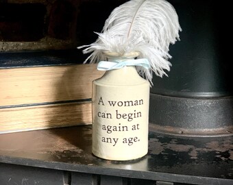 May include: A cream-colored ceramic bottle featuring the black printed text, A woman can begin again at any age. The bottle is adorned with a light blue ribbon tied around its neck and a single white ostrich feather protruding from the top opening.