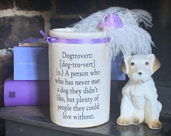 May include: A white ceramic cylindrical jar featuring black printed text that reads: Dogtrovert: [dog-tro-vert] (n.) A person who has never met a dog they didn't like, but plenty of people they could live without. A purple ribbon and white feather are attached.