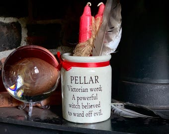 May include: A cream-colored ceramic jar with a red ribbon, containing two red candles and a large feather. The jar features the black text: PELLAR Victorian word; A powerful witch believed to ward off evil. A clear glass sphere sits to the left.