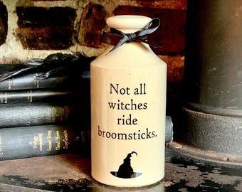 May include: A cream-colored ceramic bottle with a narrow neck and a black ribbon tied around the top. The front features the black text "Not all witches ride broomsticks." and a small black silhouette of a pointed witch hat at the bottom.
