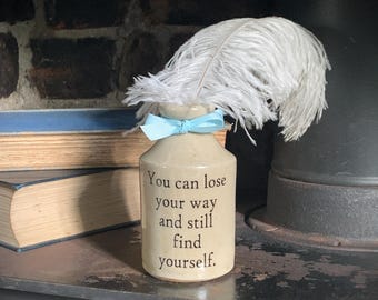 May include: A beige ceramic bottle with a light blue ribbon tied around the neck and a white ostrich feather protruding from the top. The front of the bottle features the black printed text: You can lose your way and still find yourself.