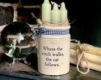 May include: A cream-colored ceramic crock featuring the black text 'Where the witch walks, the cat follows.' The crock is tied with a black and white gingham ribbon and holds three pale green candles wrapped in twine.