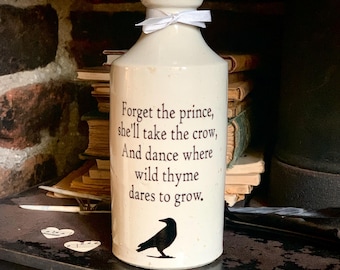 May include: Cream-colored ceramic bottle with a white bow tied around the neck. The bottle features a black crow illustration and a quote: "Forget the prince, she'll take the crow, And dance where wild thyme dares to grow."