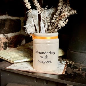 May include: A white ceramic jar with an orange ribbon around the top. The jar has the text "Meandering with purpose." written on it. Dried lavender and feathers are in the jar.