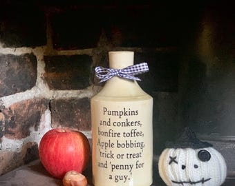 May include: A cream-colored ceramic bottle with a black and white gingham ribbon tied around the neck. The bottle features black text that reads: Pumpkins and conkers, bonfire toffee, Apple bobbing, 'trick or treat' and 'penny for a guy.'