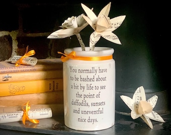 May include: A white ceramic cylindrical jar featuring the black printed text: You normally have to be bashed about a bit by life to see the point of daffodils, sunsets and uneventful nice days. It is decorated with an orange ribbon and paper flowers.