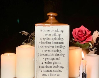 May include: A white ceramic bottle with a brown lid and a black base. The bottle has a poem printed on it that reads: "12 crones a-cackling, 11 roses wilting, 10 spiders spinning, 9 headless horsemen, 8 werewolves howling, 7 ravens cawing, 6 broomsticks dancing, 5 pentagrams! 4 restless ghosts, 3 cauldrons bubbling, 2 haunted castles, And a black cat in a dead tree."