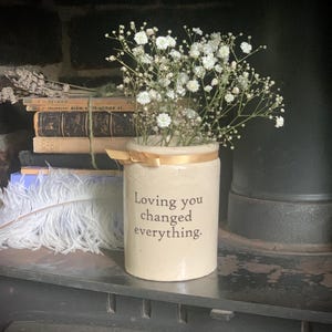 May include: A cream ceramic jar with the text "Loving you changed everything" contains white baby's breath. A gold ribbon adorns the rim. The jar is on a dark surface, with books and a feather in the background. A rustic, romantic aesthetic.