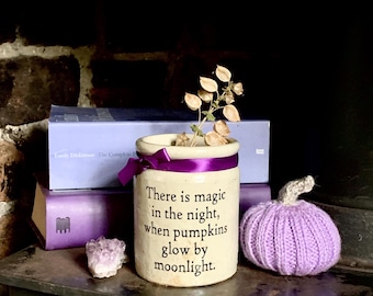 May include: A cylindrical cream-colored ceramic crock featuring a purple ribbon tied around the top and the black text: There is magic in the night, when pumpkins glow by moonlight. A small dried plant stem sits inside the crock.