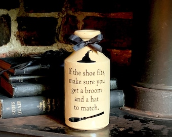 May include: A cream-colored ceramic bottle featuring a black ribbon tied around the neck. The front displays a black witch hat icon, the text "If the shoe fits, make sure you get a broom and a hat to match.", and a black broomstick graphic at the base.