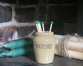 May include: A cream-colored ceramic jar with the word MATCHES printed in black lettering on the front. The jar is filled with several wooden matches that have green tips. The jar is positioned between two bundles of candles tied with twine.