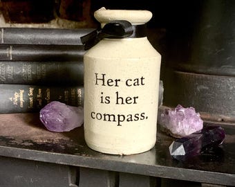 May include: A cream-colored ceramic apothecary jar with a speckled texture and a black ribbon tied around the neck. The text "Her cat is her compass." is printed in black on the front of the jar. The jar is positioned between two amethyst crystals.