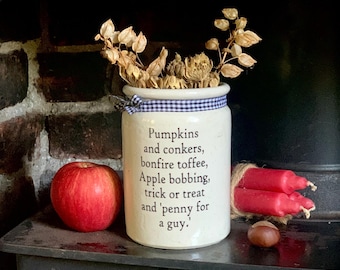 May include: A cream-colored ceramic jar featuring a blue and white checkered ribbon around the rim and black text that reads: Pumpkins and conkers, bonfire toffee, Apple bobbing, trick or treat and 'penny for a guy.' Dried foliage sits inside.