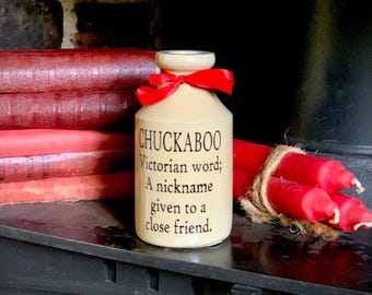 May include: A cream-colored ceramic bottle with a narrow neck and a red ribbon tied around the top. The bottle features black printed text that reads: CHUCKABOO Victorian word; A nickname given to a close friend. The bottle has a smooth finish.
