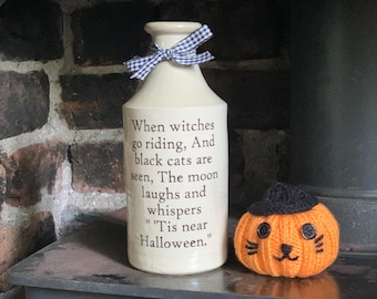 May include: A cream-colored ceramic bottle with a blue and white gingham ribbon tied around the neck, featuring the text: When witches go riding, And black cats are seen, The moon laughs and whispers 'Tis near Halloween. Beside it sits a small orange knit pumpkin.