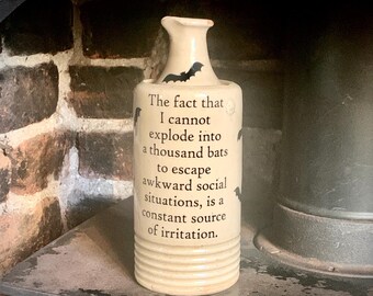May include: A cream-colored ceramic bottle with a narrow neck and ribbed base. The bottle features black bat silhouettes and the printed text: The fact that I cannot explode into a thousand bats to escape awkward social situations, is a constant source of irritation.