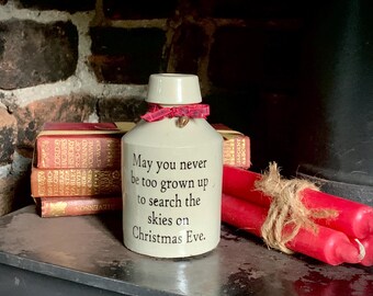 May include: A cream-colored ceramic bottle with a narrow neck and a red plaid ribbon tied around the top. The bottle features the black text: May you never be too grown up to search the skies on Christmas Eve. It is positioned near red candles.