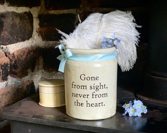 May include: A cream-colored ceramic jar featuring the black printed text 'Gone from sight, never from the heart.' The jar is adorned with a light blue ribbon tied around the top and contains a white ostrich feather and small blue flowers.