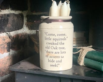 May include: A cream-colored ceramic crock filled with several white taper candles. The front of the crock features dark brown text that reads, "Come, come, little squirrels" creaked the old Oak tree, "there are lots of acorns to hide and seek!"