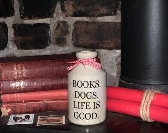 May include: A cream-colored ceramic bottle with a narrow neck and a red and white checkered ribbon tied around the top. The bottle features the black printed text: BOOKS. DOGS. LIFE IS GOOD. It is positioned among red books and red candles.