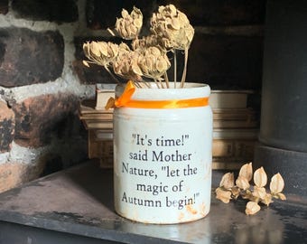 May include: A white ceramic crock featuring the black printed text, "It's time!" said Mother Nature, "let the magic of Autumn begin!" The container is accented with an orange ribbon tied around the top and holds several dried brown flower heads.