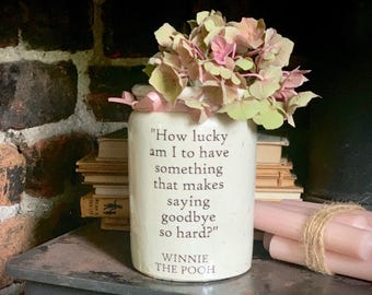 May include: A cylindrical white ceramic jar featuring the printed quote, "How lucky am I to have something that makes saying goodbye so hard?" followed by "WINNIE THE POOH" in dark lettering. The jar is filled with a cluster of pale pink hydrangea flowers.