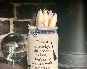 May include: A cream-colored ceramic jar filled with several off-white taper candles. The jar features a black and white gingham ribbon tied around the neck and black text that reads: The cat is snarky, the broom is fast, Don't cross a witch with spells to cast.