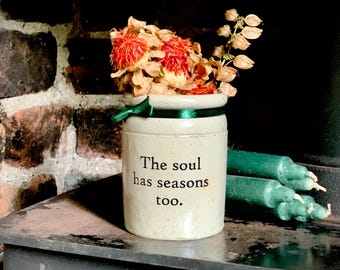 May include: A cream-colored ceramic crock with a dark green ribbon tied around the rim. The text "The soul has seasons too." is printed in black. The crock contains dried orange flowers and is next to three dark green taper candles.