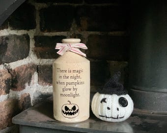 May include: A cream-colored ceramic bottle with a red and white striped ribbon tied around the neck, featuring the text "There is magic in the night, when pumpkins glow by moonlight" and a black pumpkin graphic, next to a white knit pumpkin.