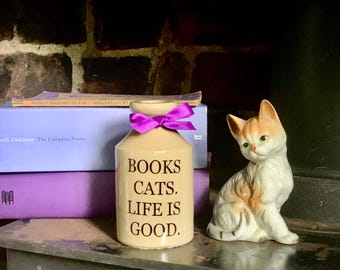 May include: A beige ceramic jar with a purple ribbon tied around the neck, featuring the black text "BOOKS CATS. LIFE IS GOOD." next to a small porcelain cat figurine with white, orange, and grey fur, positioned in front of two purple books.