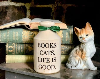 May include: A cream-colored ceramic bottle with a green ribbon tied around the neck. The text "BOOKS. CATS. LIFE IS GOOD." is printed in black on the front. The bottle is positioned next to a small, seated porcelain cat figurine.