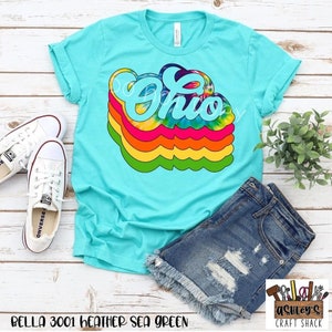 May include: A turquoise t-shirt with a tie-dye design of the word "Ohio" in rainbow colors. The shirt is laid flat on a white wooden surface with a pair of white sneakers and a pair of denim shorts.