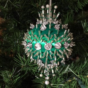 May include: A green Christmas ornament with a satin finish, adorned with clear crystals and silver bead accents. The ornament features a cluster of silver beads and crystals radiating from the center, with pearl-like beads hanging below.