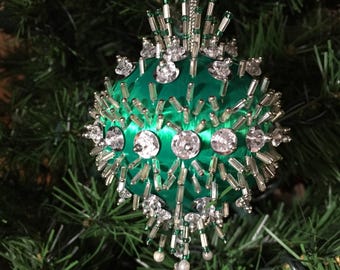 May include: A green Christmas ornament with a satin finish, adorned with clear crystals and silver bead accents. The ornament features a cluster of silver beads and crystals radiating from the center, with pearl-like beads hanging below.