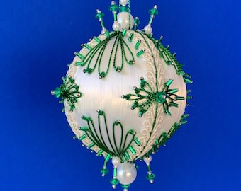 May include: A spherical ornament featuring a white satin-wrapped base decorated with intricate green beaded patterns, white braided trim, and small white pearl-like beads. The ornament hangs from a green cord and includes a dangling beaded finial.