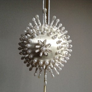 May include: A white, beaded Christmas ornament with a pearl-like finish and a silver flower design in the center.
