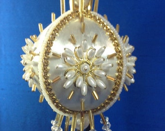 May include: A decorative, off-white ornament with gold and pearl accents. The round ornament features a central floral design and is embellished with gold beads, pearls, and crystal-like drops. It hangs from a gold chain.